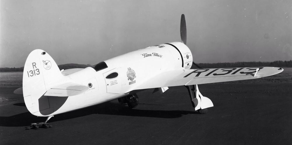 B&W photo of the Hawks-Miller HM-1 “Time Flies” racing aircraft of 1936. This aircraft (all white, but with black logos, name & registration markings) is photographed on the ground from the starboard quarter.