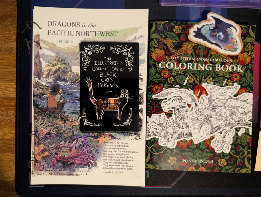 A zine titled Dragons in the Pacific Northwest, a smaller zine titled The Illustrated Collection of Black Cats Musings, a coloring book titled Itty Bitty Seasonal Dragons, and a dragon curled around a pearl sticker - all drawn by Indui.