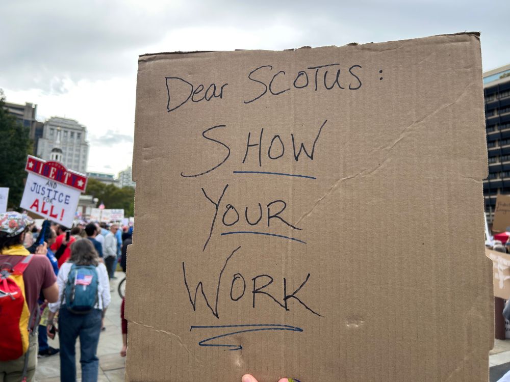 A sign that says “Dear SCOTUS: SHOW YOUR WORK”, held up at a rally with Independence Hall in the background 