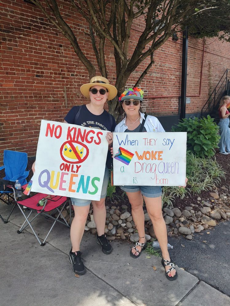 2 women holding protest signs. One says, "No kings only Queens" with a crossed of crown. The other states, "when they say woke a drag queen is born" with the progress flag.