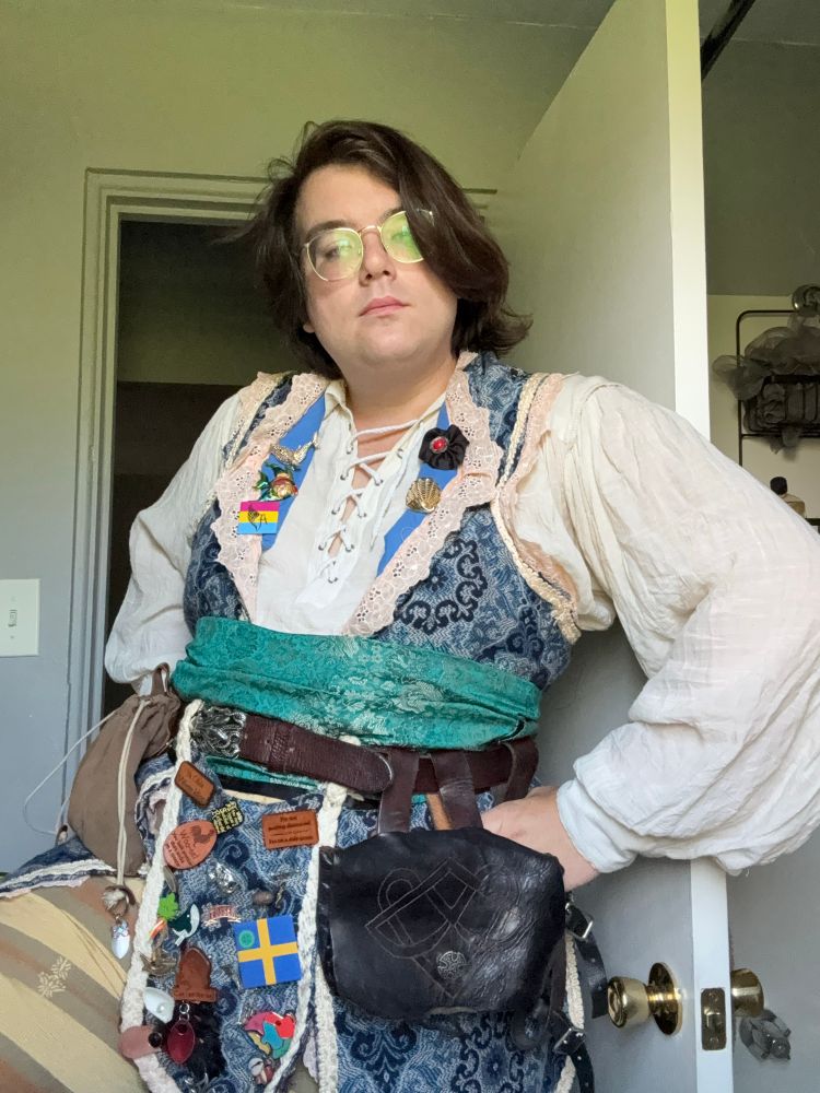 Zel is wearing her renaissance faire garb, which consists of a flowy linen shirt, a blue and pink vest, teal sash and kraken buckle belt, as well as pouches hanging from the belt and beige/brown pants
