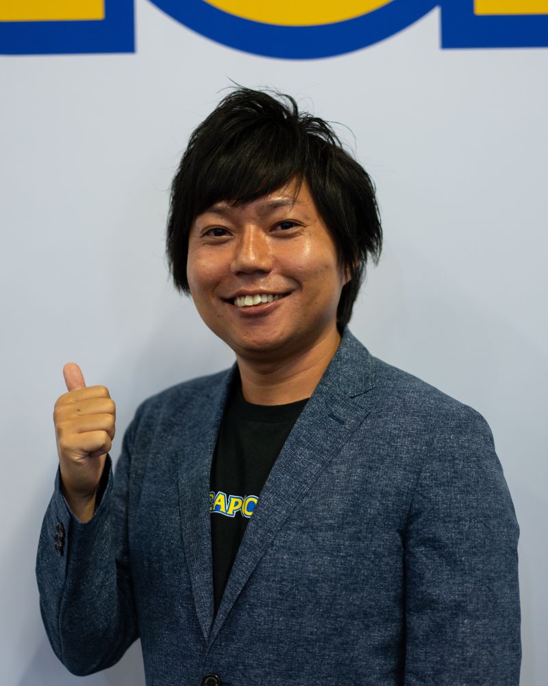 A portrait of Satoru Nihei, Director of Onimusha: Way of the Sword standing in front of the Capcom booth at Gamescom. He smiles into the camera while giving a thumbs up.