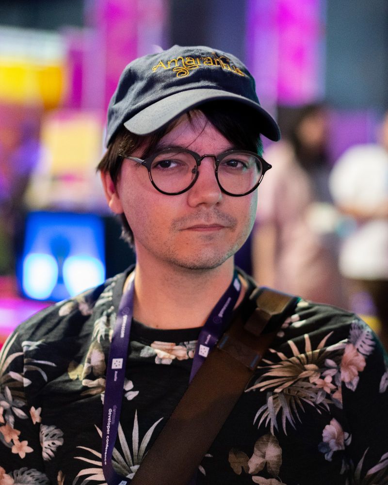 A portrait of Game Trailer Maker Gary Kings. The background is colorful and dominated by the color purple. He is wearing a shirt displaying various plants. His cap and glasses in combination with his eyes are exuding confidence as if he knows the picture will come out well. 