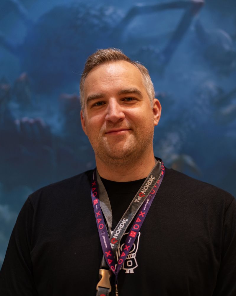 A portrait of Stefan Mohr, Producer at THQ Nordic. He is standing in front of a blue background wearing a black shirt with a small drawing on it as well as multiple lanyards around his neck. One clearly reads THQ Nordic. He is displaying a confident smile into the camera. 