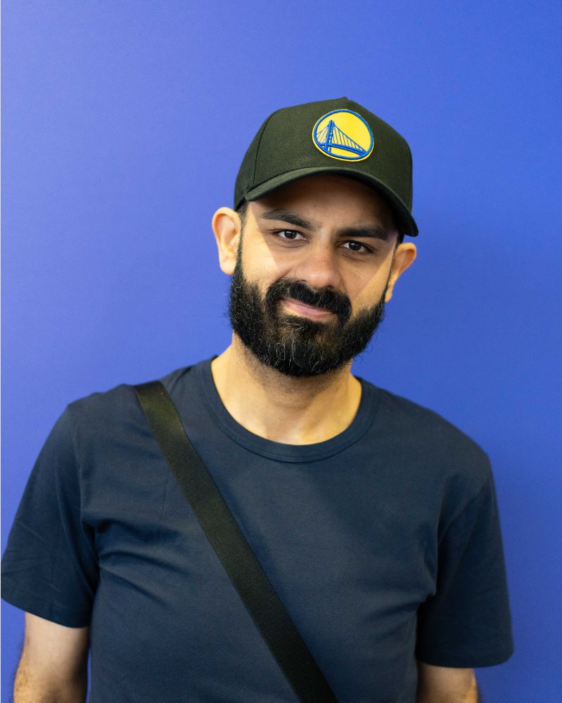 A portrait of Tamoor Hussain, Managing Editor at Gamespot. He is standing in front of a blue booth, wearing a black cap with a bridge on it. He elegantly smirks into the camera. 