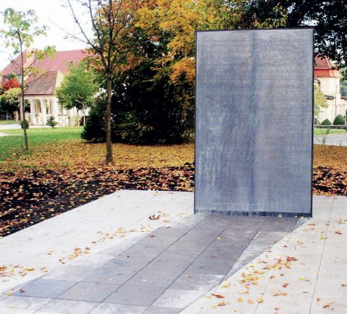 Das Bild zeigt ein Denkmal in einer parkähnlichen Umgebung. Im Mittelpunkt steht eine schlichte, rechteckige, vertikale Stele aus grauem Metall oder Stein. Vor der Stele verläuft auf dem Boden ein schmaler Streifen aus dem gleichen Material, der wie ein Schatten wirkt. Die Umgebung ist herbstlich: Der Boden ist mit bunten Blättern bedeckt, und im Hintergrund stehen Bäume mit teils gelbem, teils grünem Laub. Hinter den Bäumen sind Gebäude mit roten Dächern und gelblichen Fassaden zu sehen, die an historische Architektur erinnern. Das Denkmal wirkt zurückhaltend und schlicht, was möglicherweise der Würde des Themas Rechnung trägt, für das es errichtet wurde.
