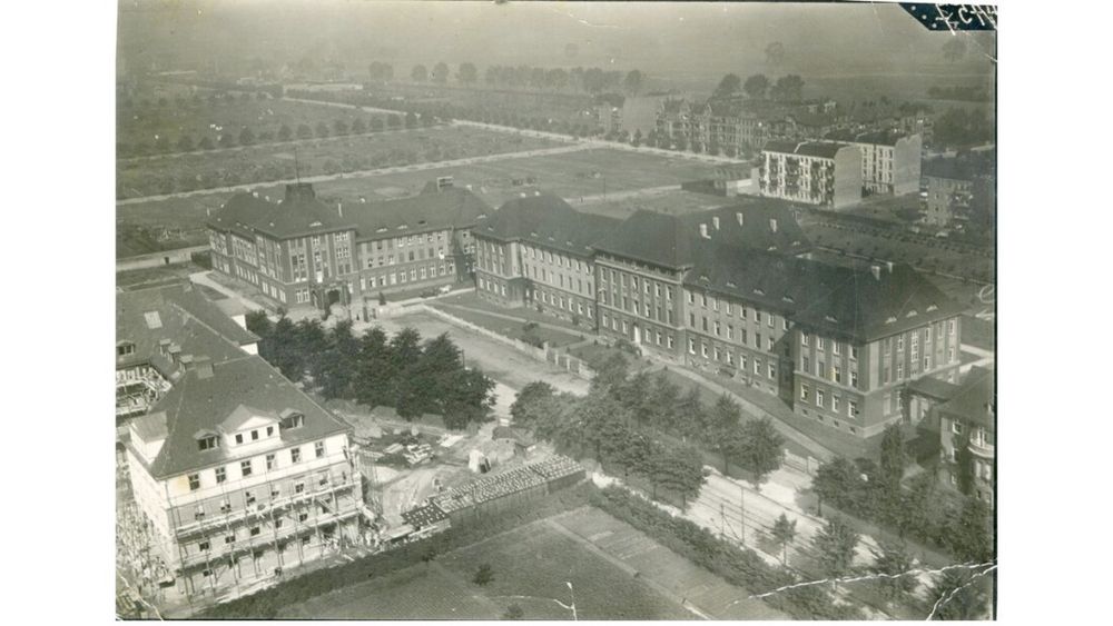 Das Bild zeigt eine historische, schwarz-weiße Luftaufnahme eines großen Gebäudekomplexes, das wie eine Klinik oder eine Schule aussieht. Es handelt sich um eine mehrstöckige Anlage im traditionellen Baustil mit dunklen, schrägen Dächern und zahlreichen Fenstern.

Der Komplex besteht aus mehreren Flügeln, die um Innenhöfe angeordnet sind, mit breiten, asphaltierten Wegen und Bäumen in der Umgebung. Ein Bereich scheint gerade im Bau zu sein – im Vordergrund sind Baugerüste und Baumaterialien zu erkennen.

Im Hintergrund sieht man eine weitläufige, offene Landschaft mit vereinzelten Gebäuden, Straßen und Bäumen. Insgesamt vermittelt das Bild eine ruhige, städtische Szenerie aus einer früheren Epoche.