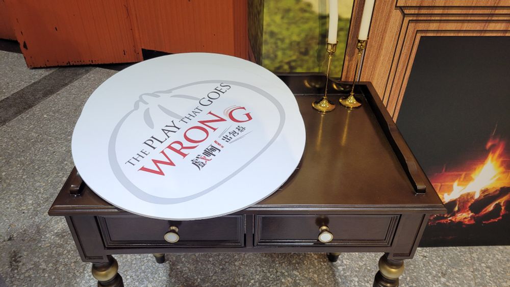 Selfie props for The Play That Goes Wrong, Taiwan edition, including a large board depicting the name of the play on a table with two candles sticks