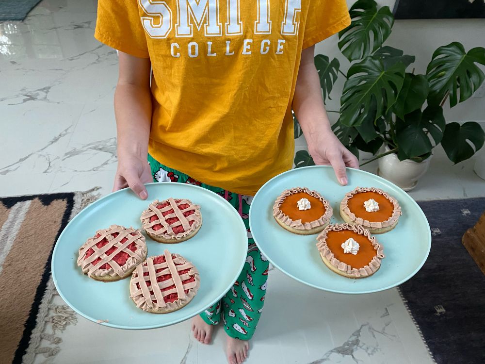 A teenager wearing Christmas themed PJ pants and a Smith College tshirt (whose face is not visible) holds two plates. One has three cookies that look like miniature cherry pies with cross hatched frosting that looks like pie crust. The cookies on the other plate look like miniature pumpkin pies with whipped cream topping in the middle.