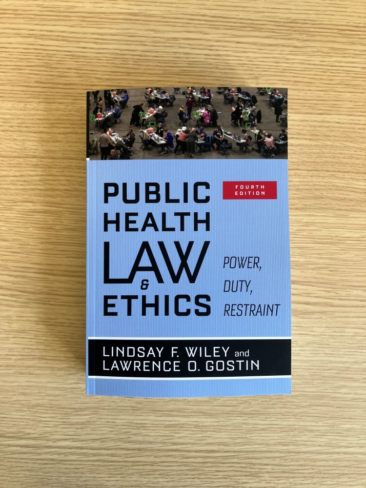 A thick paperback book rests on a wooden table, presumably in my editor's office, where it awaits being placed into a box and shipped to me. The cover is mostly light blue and reads "Public Health Law & Ethics: Power, Duty, Restraint by Lindsay F. Wiley and Lawrence O. Gostin. Along the top edge of the cover is a photograph providing a bird's eye view of about 100 people receiving and administering vaccinations en mass in a large open room. 