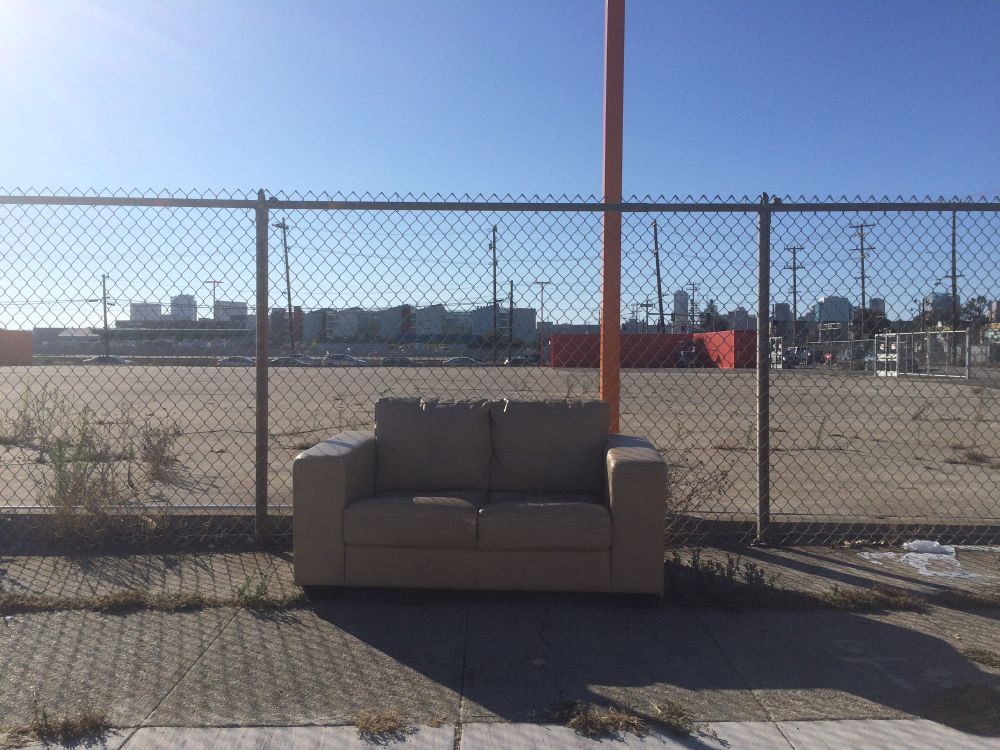 A couch abandoned in front of an empty fenced in lot