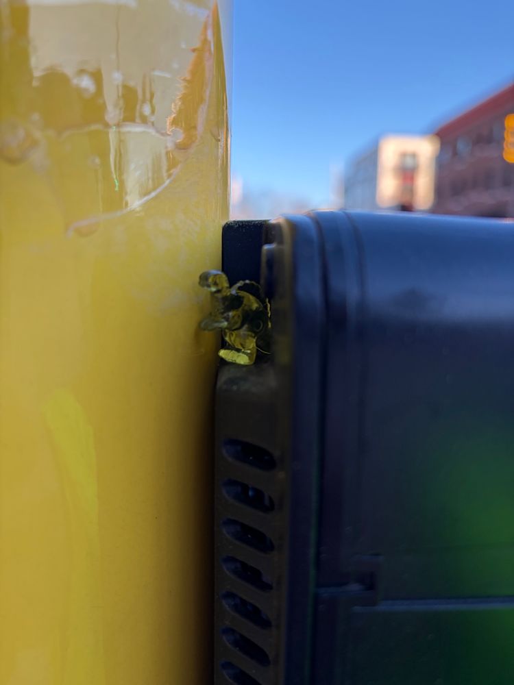 A small yellow plastic dinosaur is tucked tightly into the narrow gap between a glossy yellow metal pole and a black box. Only part of the tiny figure is visible from the cramped space.