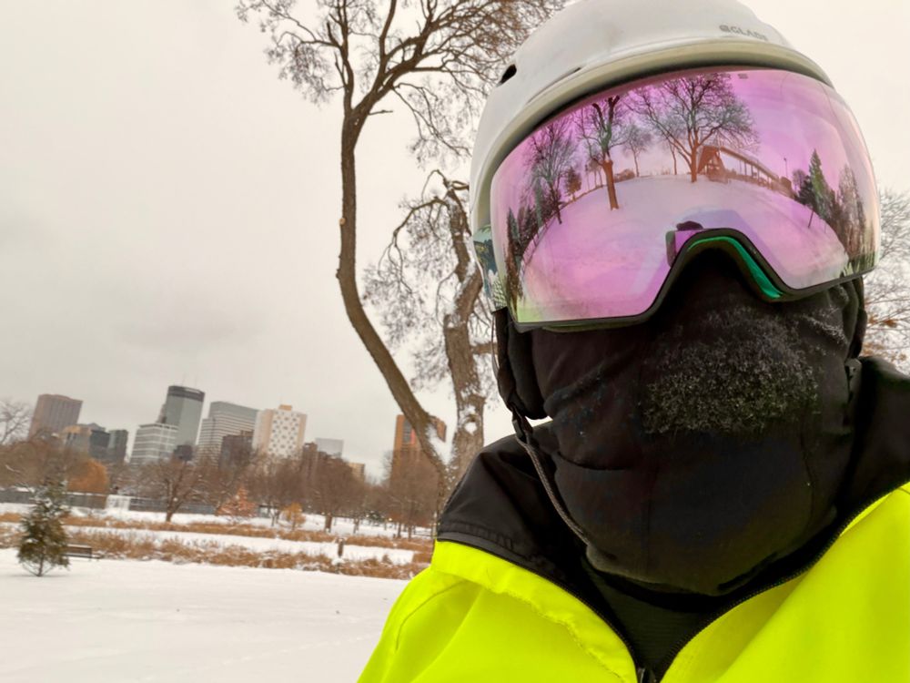 A person bundled for winter biking takes a selfie outdoors. They wear a white helmet, reflective pink ski goggles, and a black face covering with frost on it. Behind them is a snowy park with bare trees and the Minneapolis skyline under a gray winter sky reflected in their goggles.