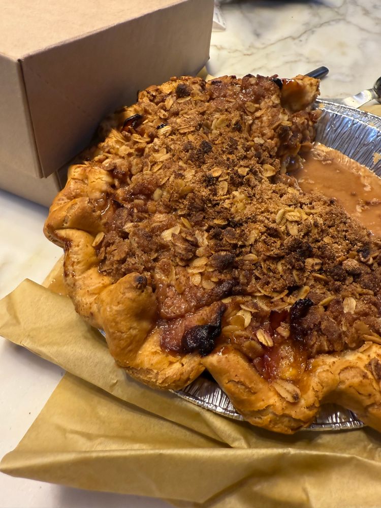 A rustic peach pie in an aluminum tin, topped with a chunky oat–brown sugar crumble; flaky crimped crust with some caramelized peach filling oozing out, set on parchment on a marble counter with a cardboard pie box in the background
