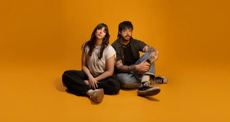 Austin and Taryn Duffy, the brother and sister team at the core of the band, sit against an orange background. Austin has short dark hair, glasses, facial hair and tattoos. Taryn has long dark hair. Both are wearing jeans and t-shirts.