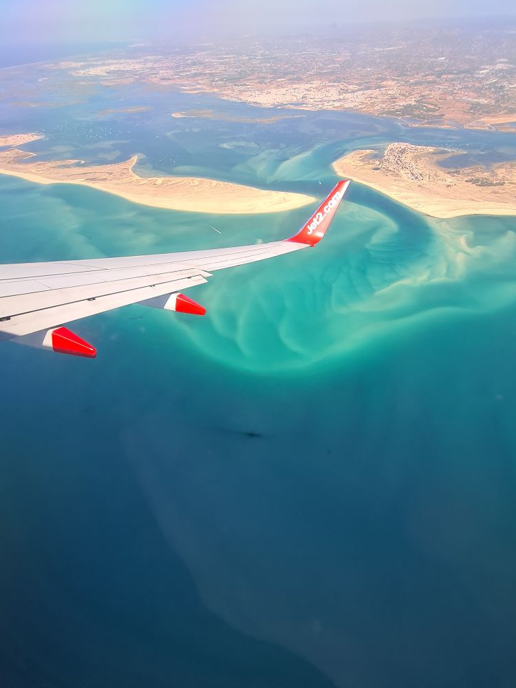 Picture of beautiful crystal clear sea with beach islands in the background, leading to the south of Portugal. In the foreground there is a wing of a Jet2 plane and in the sea there is a shipwreck. 