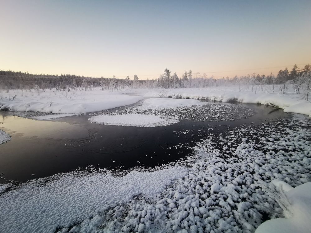 A dark river, partially frozen over with patches of snow and ice, winds through a snow-covered landscape. The banks of the river are lined with snow, and a distant line of snow-covered trees is visible under a pale, twilight sky.