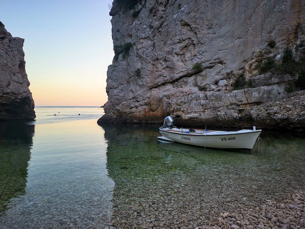Ein Boot befindet sich in einem kleinen, felsigen Tal am Meer. Das Boot ist weiß mit einem kleinen Außenbordmotor am Heck. Die Felsen an beiden Seiten sind hoch und dunkel, und das Wasser dazwischen ist klar und reflektiert das Licht. Der Himmel ist blau und grau. Auf der Seite des Bootes steht in schwarzen Buchstaben "STINIVA". Auf der Rückseite des Bootes steht in schwarzen Buchstaben "VS 460".