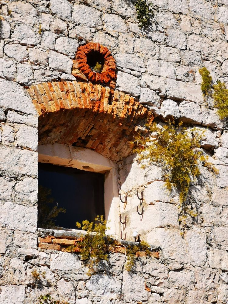 The image shows a close-up view of an old stone wall with a window. The wall is constructed from irregularly shaped, light-colored stones, giving it a rugged appearance. The window is rectangular with a brick arch above it, featuring a circular brick feature at the top. The window is partially open, revealing a dark interior. The wall shows signs of age, with some plants growing through the cracks, adding a touch of greenery. Metal hooks are visible on the wall, suggesting it may have been used for hanging items. The overall texture of the wall is rough, with a mix of stone and brick elements, and the lighting suggests it is a sunny day.