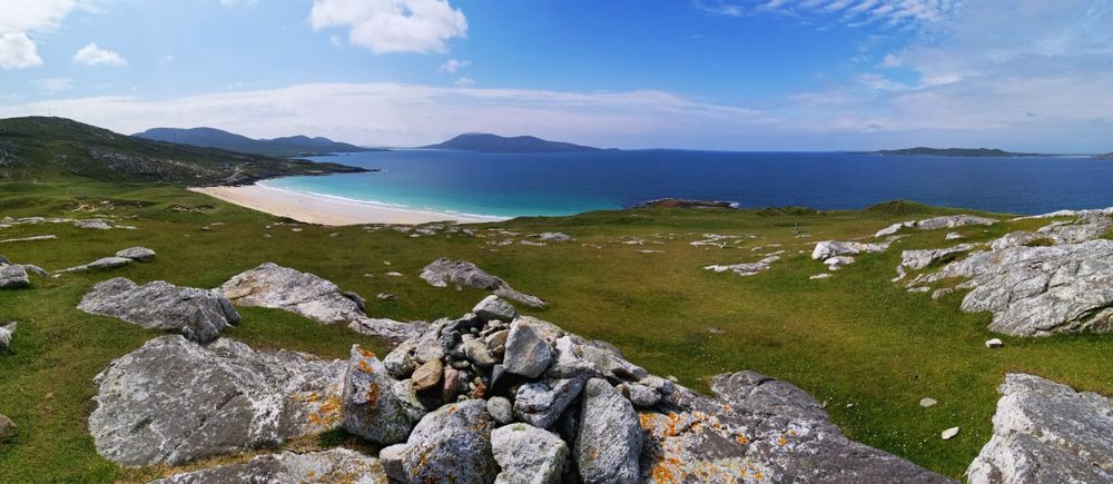 The image depicts a serene coastal landscape. In the foreground, there is a grassy area with scattered large, gray rocks, some covered with patches of orange lichen. The middle ground features a sandy beach with turquoise waters, bordered by gentle hills. In the background, the ocean extends to the horizon, with distant islands visible. The sky is mostly clear with a few scattered clouds, suggesting a calm day. The overall scene is tranquil and picturesque, showcasing the natural beauty of the coastal region.
