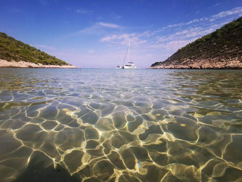 A clear, shallow body of water with a sandy bottom is visible. The water is textured by the light shining through it. In the distance, a sailboat is anchored between two small, green, rocky hills under a bright blue sky with a few clouds