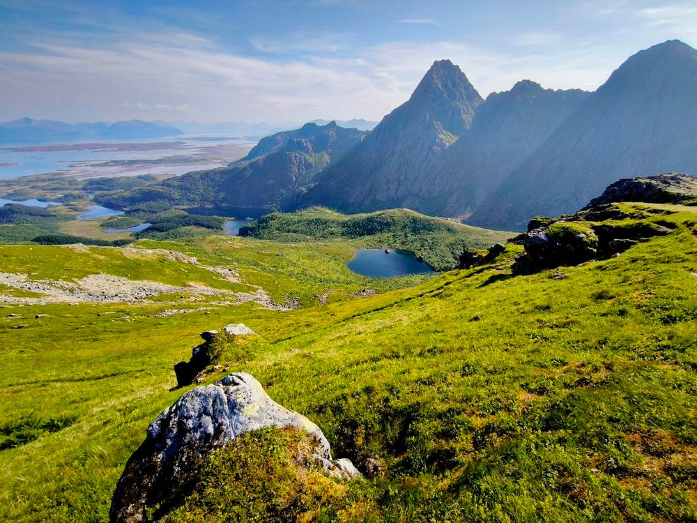 The image depicts a stunning mountainous landscape under a clear blue sky with scattered clouds. In the foreground, there is a grassy hillside with patches of green grass and a large, moss-covered rock. The middle ground features a serene lake nestled in a valley, surrounded by lush greenery. The background showcases towering, rugged mountains with sharp peaks, creating a dramatic contrast against the sky. The mountains are partially shrouded in mist, adding depth to the scene. The overall color palette is vibrant, with the greens of the grass and trees, the blues of the sky and water, and the grays and browns of the rocky terrain. The image captures the natural beauty and tranquility of the landscape, with no visible human presence or man-made structures.