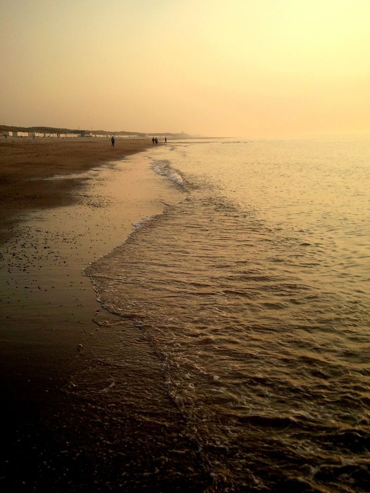 A long shot of a sandy beach at sunset. The sand is dark brown and wet, with small waves gently lapping the shore. A few people are visible in the distance, walking along the beach. In the background, there is a line of low buildings or beach huts. The sky is a pale, hazy yellow-orange.