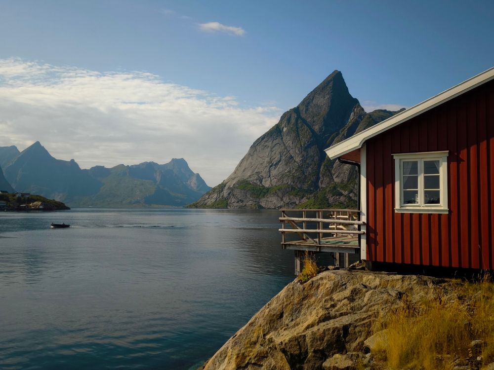 The image depicts a serene landscape featuring a red wooden house with white trim, situated on a rocky outcrop by a calm body of water. The house has a single window and a small wooden deck with a railing, extending over the water. In the background, towering mountains with steep, rugged peaks rise dramatically against a partly cloudy sky. A small boat is visible on the water, adding a sense of scale and tranquility to the scene. The overall atmosphere is peaceful and picturesque, with the natural beauty of the landscape complementing the rustic charm of the house.