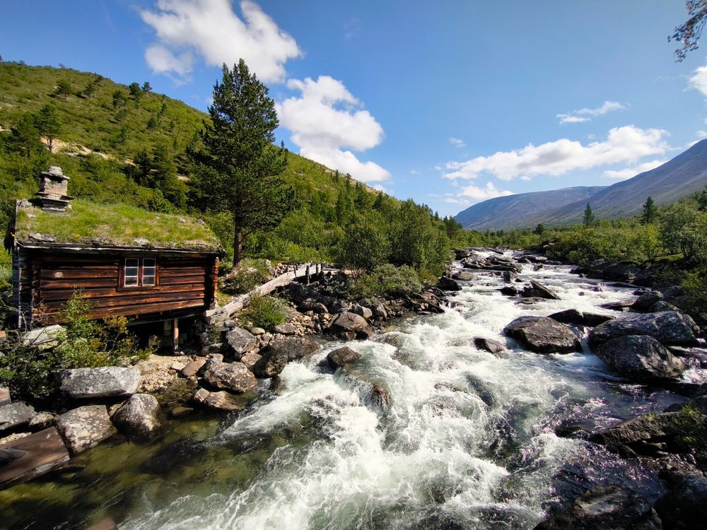A dark brown wooden cabin with a grass roof sits on the bank of a swiftly flowing river. The river is filled with rocks and rapids. Behind the cabin and river, a green hillside rises to meet a mountain range under a bright blue sky with fluffy white clouds.