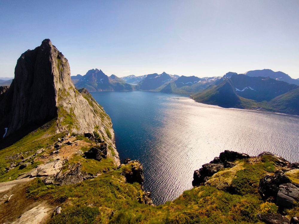 The image depicts a breathtaking landscape featuring a fjord surrounded by towering mountains. In the foreground, a steep, rocky cliff with patches of green vegetation rises sharply from the ground, leading to a rugged terrain. The cliff's surface is rugged, with visible rock formations and sparse vegetation. The middle ground showcases the calm, reflective surface of the fjord, with sunlight creating a shimmering effect on the water. The fjord is flanked by a series of mountains, some with snow-capped peaks, indicating a high altitude. The mountains in the background are more distant and less detailed, creating a sense of depth. The sky is clear and blue, suggesting a sunny day. The overall scene is serene and untouched, highlighting the natural beauty of the landscape.