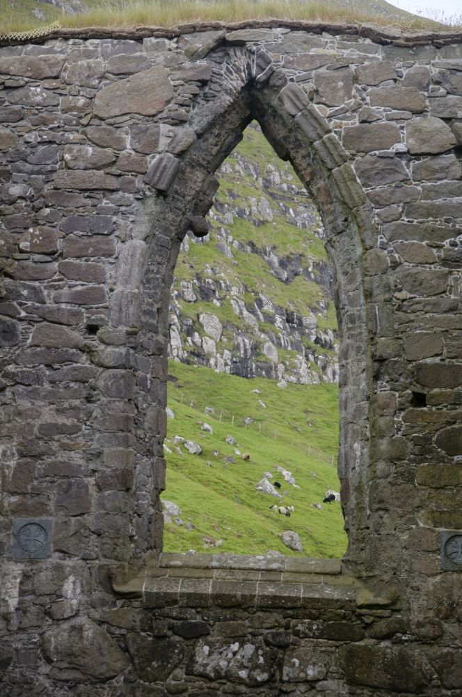 The image shows the ruins of a stone structure with a large, arched window opening. The window is framed by weathered, irregularly shaped stones, some of which are moss-covered, indicating age and exposure to the elements. The arch is pointed, typical of Gothic architectural style, and the stonework is rough and uneven, suggesting it is part of an old building, possibly a church or castle. The window provides a view of a grassy hillside with scattered rocks and a few grazing sheep. The hillside is steep and rugged, with patches of grass and exposed rock, and the sky is overcast, giving the scene a muted, natural light. The overall atmosphere is one of historical significance and natural beauty.