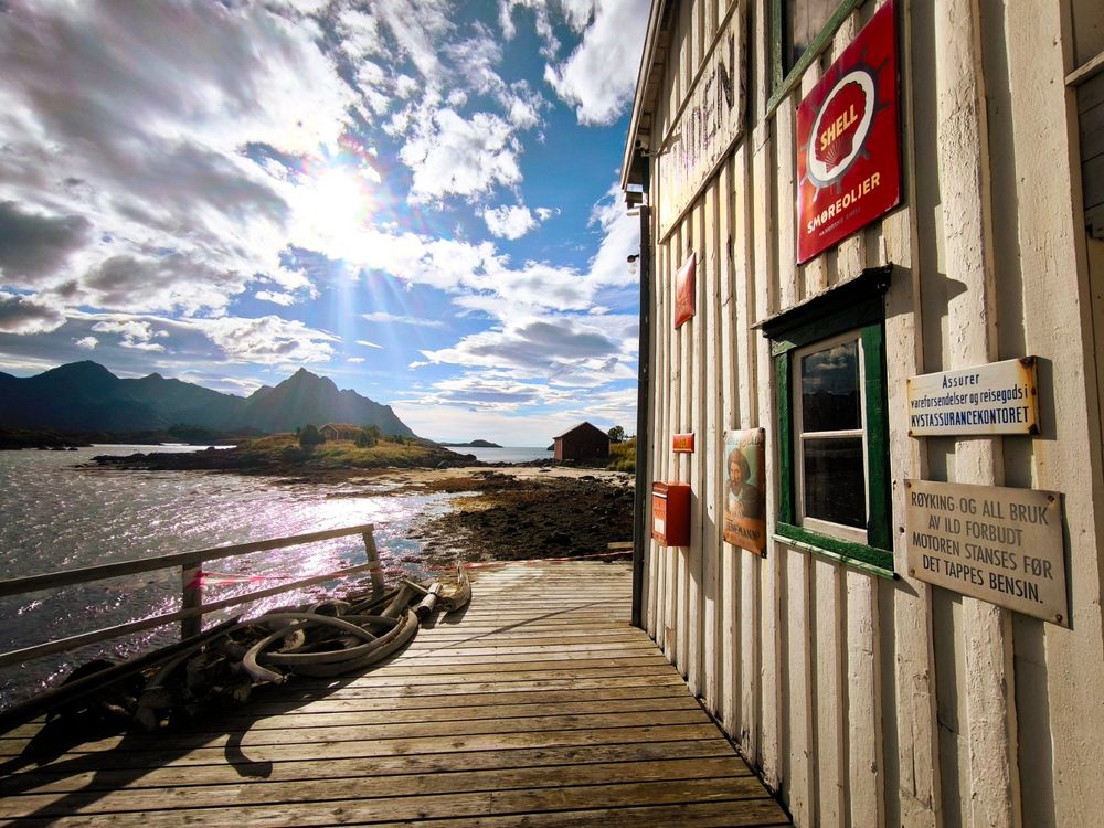 The image depicts a serene coastal scene with a wooden building on the right side, featuring a Shell sign and a red sign with the word "Smorelier." The building has a window with green trim and a small red mailbox. A coiled rope is visible on the wooden deck in the foreground. The background showcases a calm body of water with a small island and mountains under a partly cloudy sky, with the sun shining through the clouds. There are two signs on the building: one in Norwegian stating "Assurer" and another warning about the prohibition of smoking and the consequences of spilling fuel. The overall atmosphere is peaceful and picturesque, with natural elements and man-made structures harmoniously coexisting.