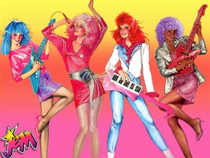 4 members of Jem and the Holograms from left to right: Aja, Jem, Kimber, Shana. Aja and Shana are playing guitar. Kimber is playing keytar, and Jem is singing into microphone. Aja has blue hair and wears a pink shirt and purple miniskirt. Jem has pink hair and wears a pink minidress with fringes belt. Kimber has red hair and wears blue capris, white blazer, and pink blouse underneath with cuffs that peek through end of sleeves. Shana has purple Afro and wears purple dress with silver blazer on top. The background is yellow, orange, purple color gradient. Jem logo is in bottom left corner.