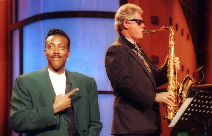 Still of 90s Bill Clinton playing the saxophone in sunglasses and a suit,  next to Arsinio Hall in a large, extremely 90s green suit pointing at him and smiling
