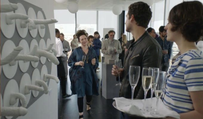 Screencap from the show Fleabag of a wall of sculputure penises, with the artist walking towards two people with champagne looking at the wall confusedly.