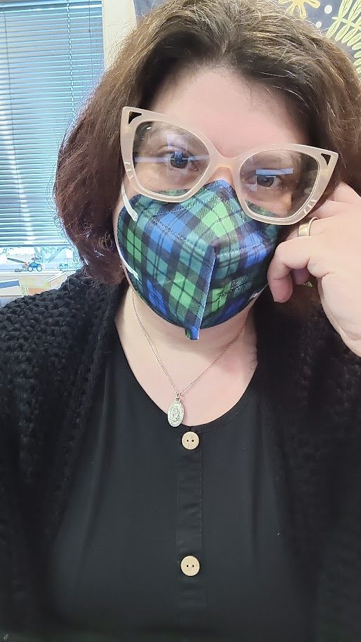 A person with brown hair and cream colored cat eye glasses rests their cheek on their hand. They are wearing a black dress with a black cardigan over it, and their kn95 mask is blue, green, and black plaid.
