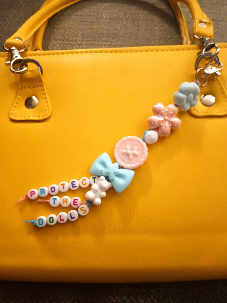 A yellow leather clutch with an acrylic beaded bag charm in the trans pride colors, with alphabet beads at the bottom that read 'protect the dolls'.