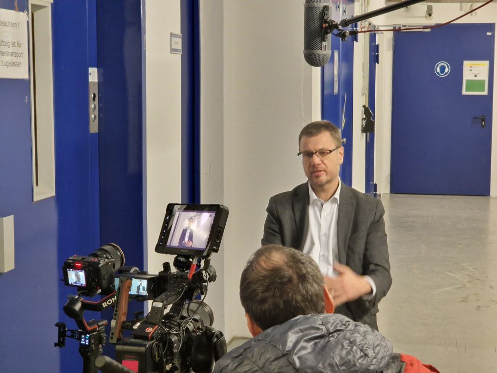 Mann sitzt ineinem Flur mit mehreren blauen Türen auf einem Stuhl und wird von zwei Kameras gefilmt, im Vordergrund der Hinterkopf eines weiteren Manns mit Kapuze. 