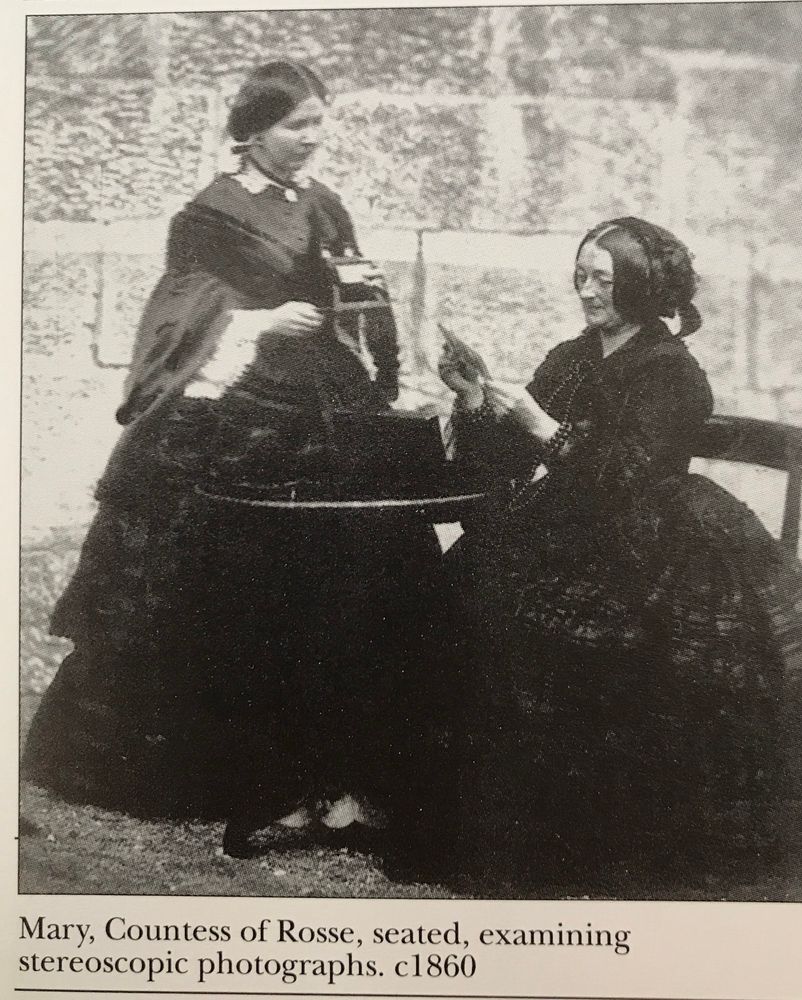 Reproduction of a 19th c photograph of two women, both in voluminous dark dresses, the elder sitting at a table looking at a stereoview, while her younger companion holds a stereoviewer. The caption reads ‘Mary, Countess of Rosse, seated, examining stereoscopic photographs, c.1860’