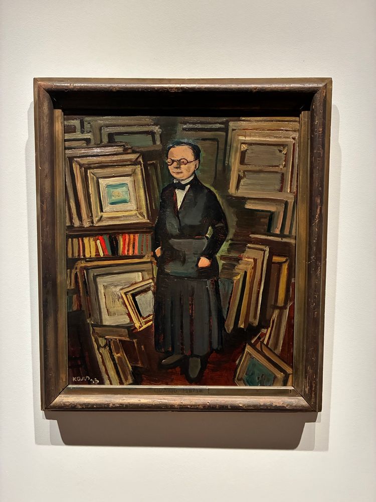 Painting by Georges Kars, showing Berthe dressed in somber clothing, wearing glasses, surrounded by stacks of frames and paintings
