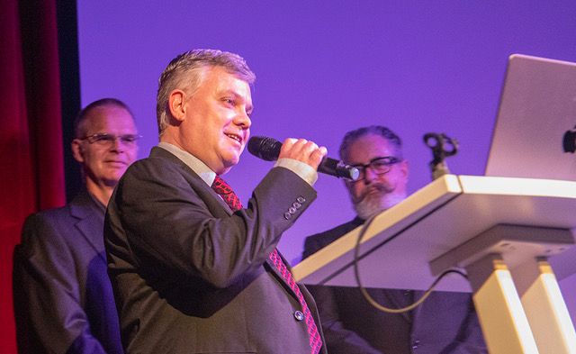 Judge Richard Todman stands at a white lectern holding a microphone. Behind him is a man with a huge twirly moustache, Roland Dunn, and a tall, bespectacled gentleman Tim Fox. Behind them is a blue cinema screen and on the left is a red curtain. 