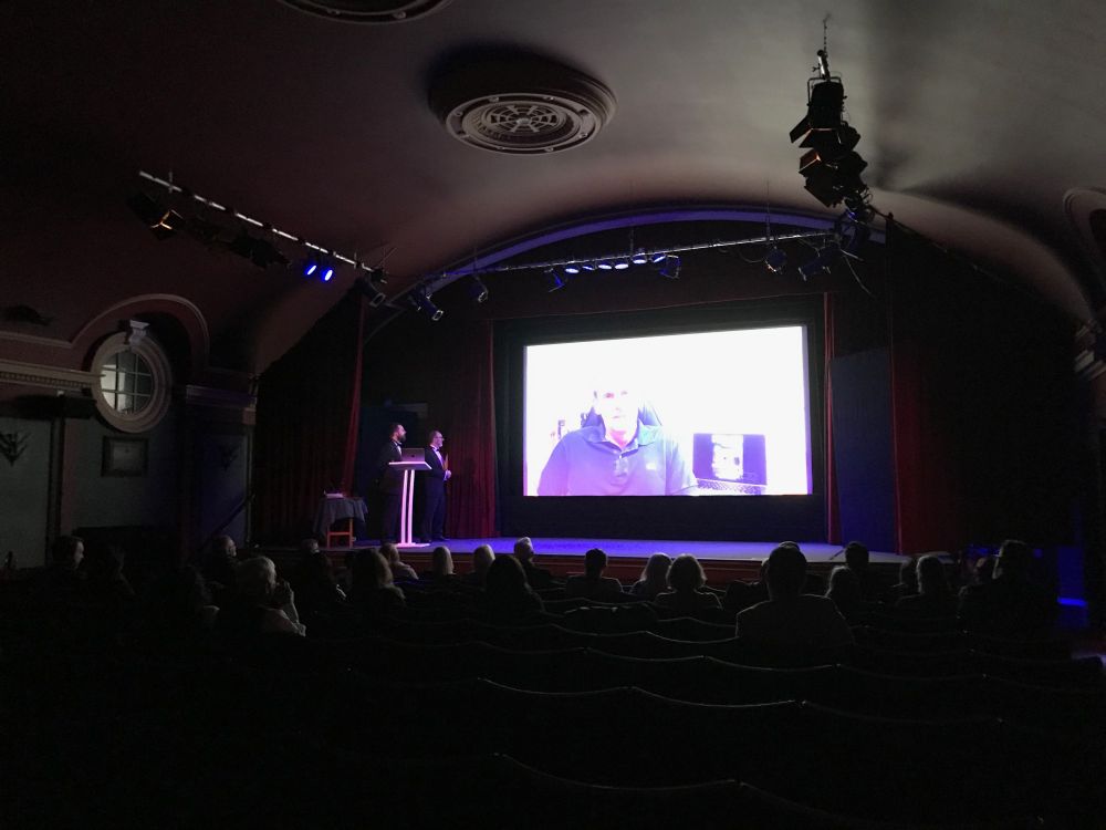 The Hailsham cinema in darkness with an audience sitting and watching a video of a man giving an acceptance speech. Two presenters stand to the left of the stage behind a lectern watching the screen. 