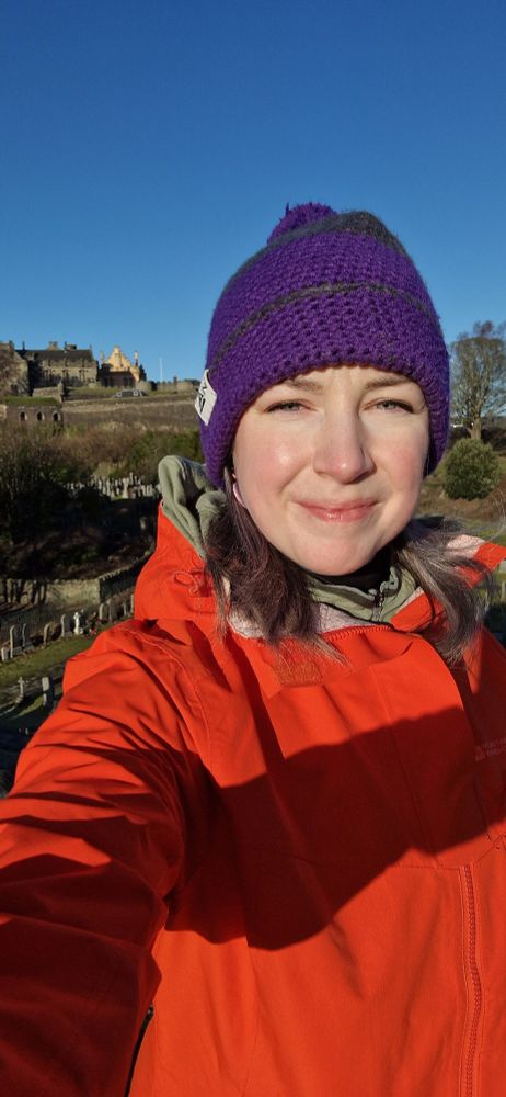 Selfie of woman in purple beanie hat and orange coat, clear blue sky in background