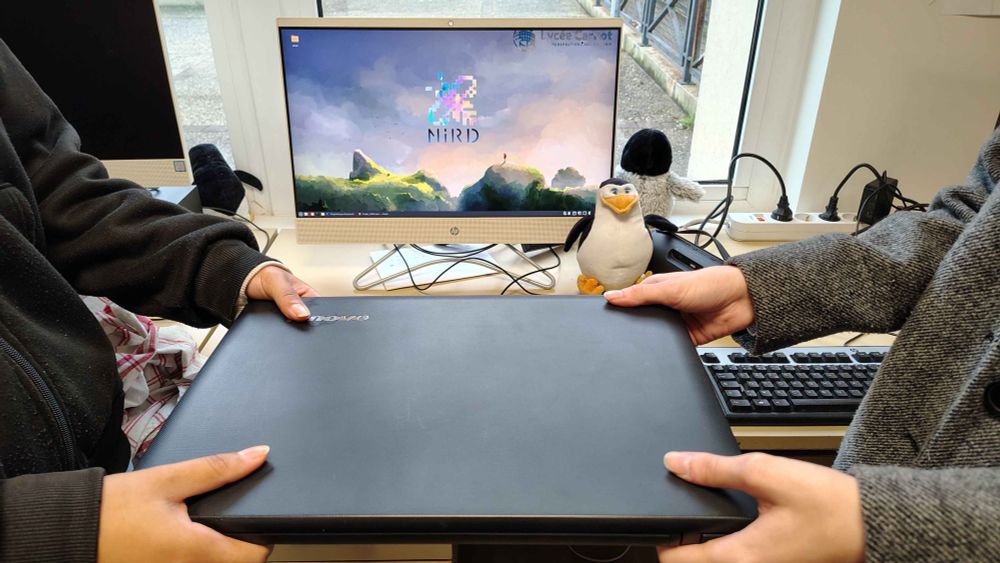 Un(e) lycéen(e) remet un Pc recondtionné #Linux à un(e) autre.
Un Pcs fixes sous Linux intégré au réseau est visible dans le fond de l'image, fond d'écran logo NIRD.