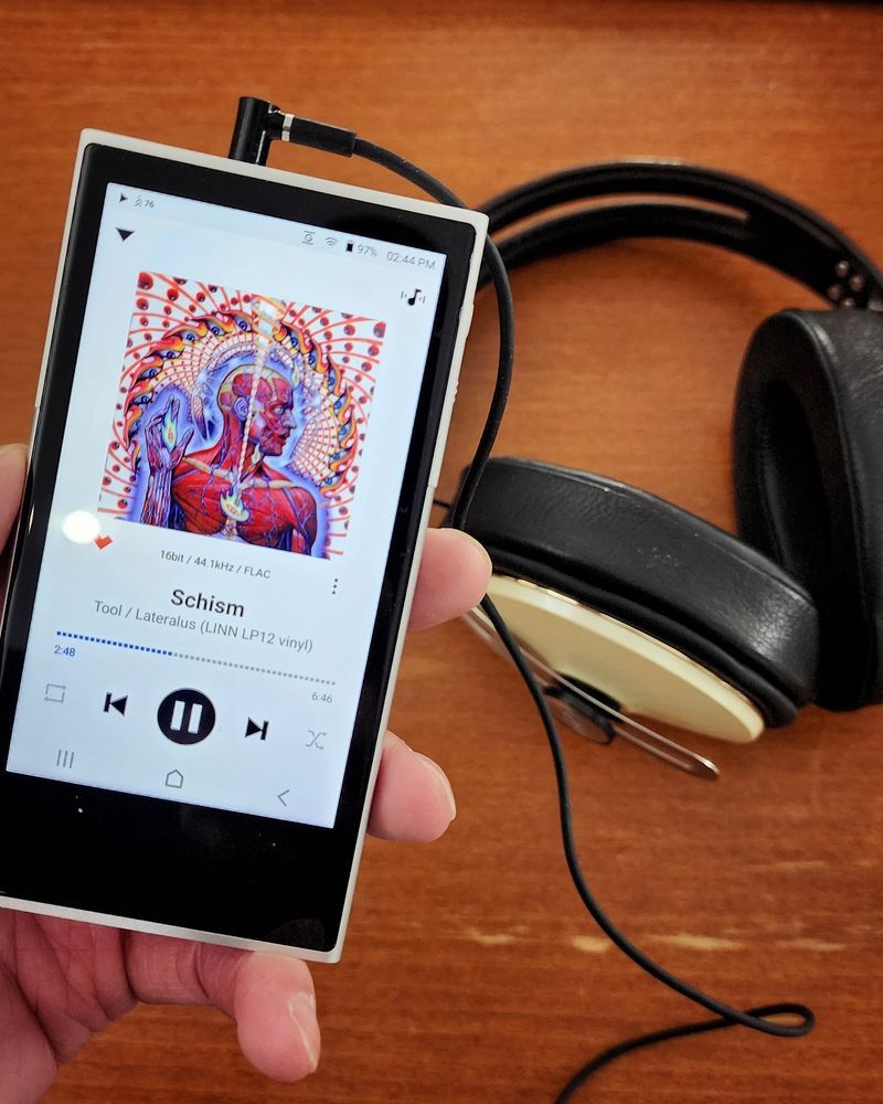 A close up look of the Activo P1 DAP with an over ears headphones in the background. Schism by Tool is shown playing on the native music app on the DAP.