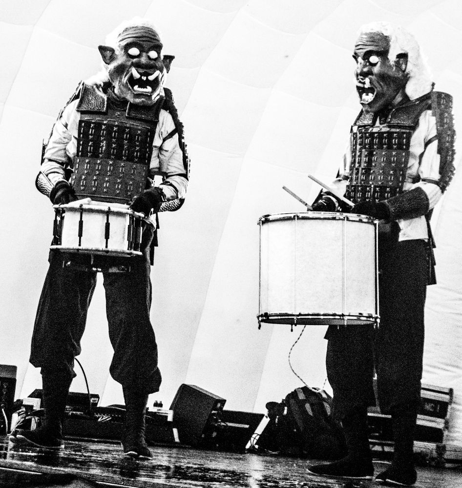 black and white photograph of of 2 people in strange oversized masks, with tusks, big white eyes and a kind of samurai clothing costume, playing percussion drums