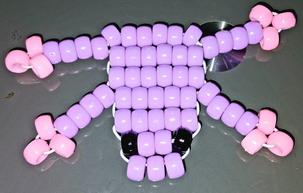 An image of a kandi frog, made using a ladder stitch. The body of the frog is light purple in colour, with black eyes, and the feet (three toes each, very important) are light pink, to contrast with the purple. 