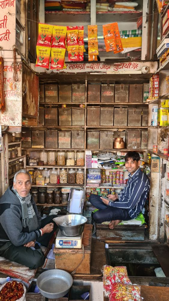 A narrow shopfront with two men sitting on the floor, the back wall filled with shelves with boxes.
