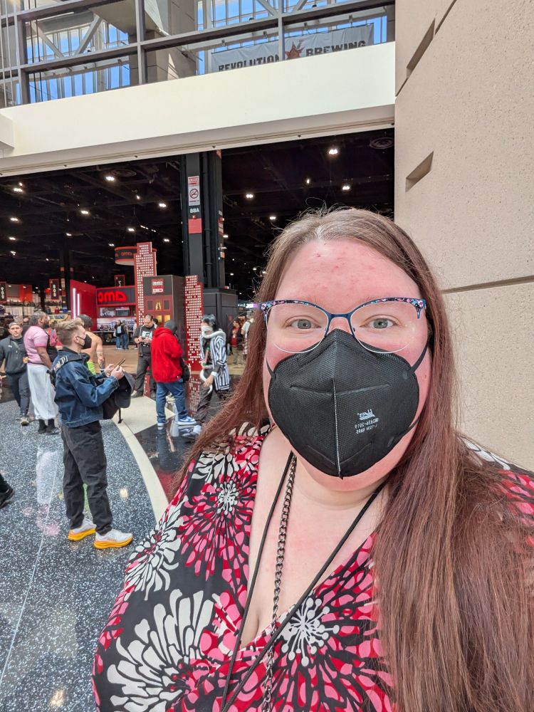A selfie of a white woman in a black face mask, with a con floor behind her.
