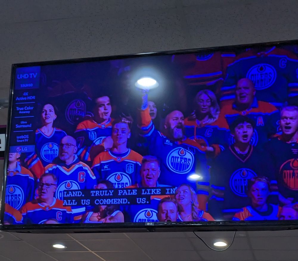A photo of a TV showing an anthem singer in a set of Oilers fans. The closed captioning reads "land. Truly pale like in all us commend. Us."